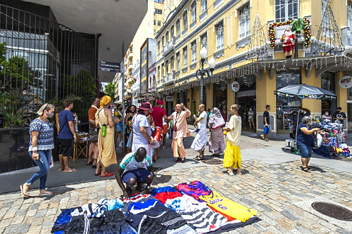 Vendedor ambulante de roupas esportivas e ao fundo grupo Hare Krishna em rua comercial no centro da cidade - 